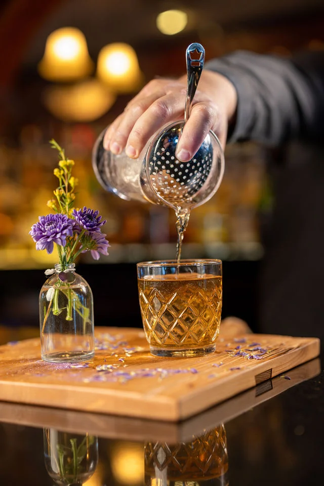 Bartender pouring a golden cocktail over ice in a cut-glass tumbler beside purple flowers at Mark’s Prime